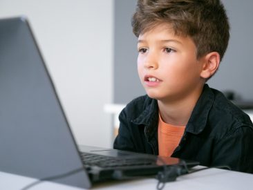 Caucasian boy sitting at table in classroom, reading text on screen or watching video presentation. Concentrated pupil studying in computer school. Knowledge and digital education concept